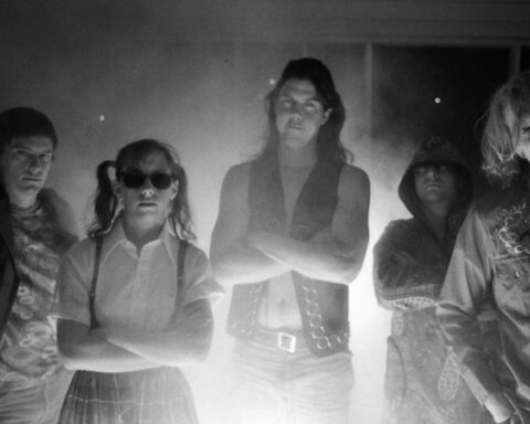 A black and white photo of the band Butthole Surfers. Five people stand looking at the camera with their arms crossed. There is a lens flare in the middle of the photo.