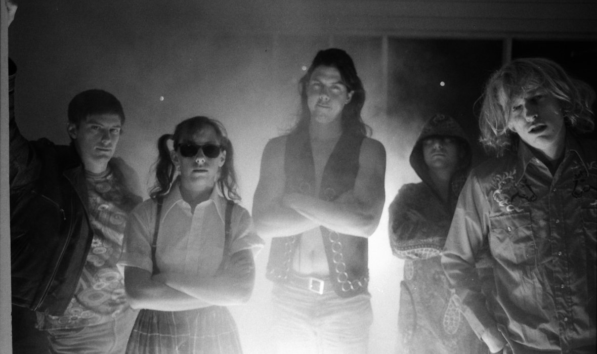 A black and white photo of the band Butthole Surfers. Five people stand looking at the camera with their arms crossed. There is a lens flare in the middle of the photo.