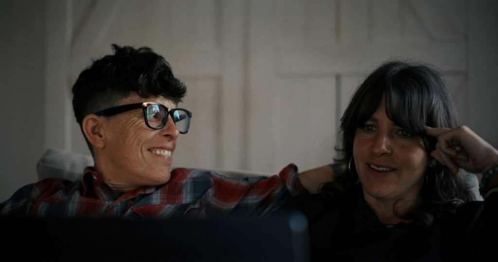 Poets Andrea Gibson and Megan Falley sit together on a couch in the documentary Come See Me in the Good Light. They are seen in a medium shot.