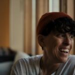 Poet Andrea Gibson is seen in close-up. They are laughing, and are a white person with short brown hair, wearing a white t-shirt and red toque.