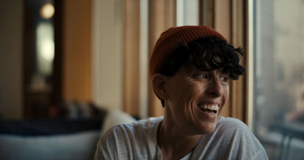 Poet Andrea Gibson is seen in close-up. They are laughing, and are a white person with short brown hair, wearing a white t-shirt and red toque.