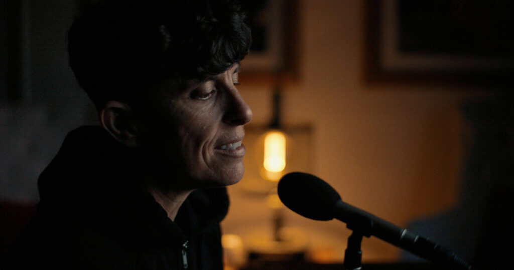 Poet Andrea Gibson is seated at a microphone. They are in a darkened room with soft light behind them.