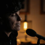 Poet Andrea Gibson is seated at a microphone. They are in a darkened room with soft light behind them.