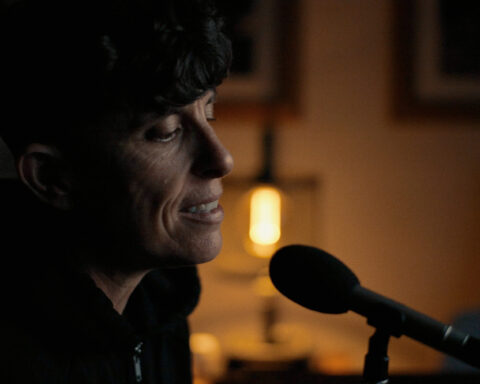 Poet Andrea Gibson is seated at a microphone. They are in a darkened room with soft light behind them.