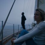 A blonde woman sits on a sailboat with her hands clasped on her knees while looking back towards her father standing at the end of the boat. It is a dark and cloudy day.