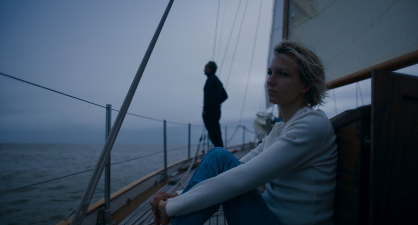 A blonde woman sits on a sailboat with her hands clasped on her knees while looking back towards her father standing at the end of the boat. It is a dark and cloudy day.