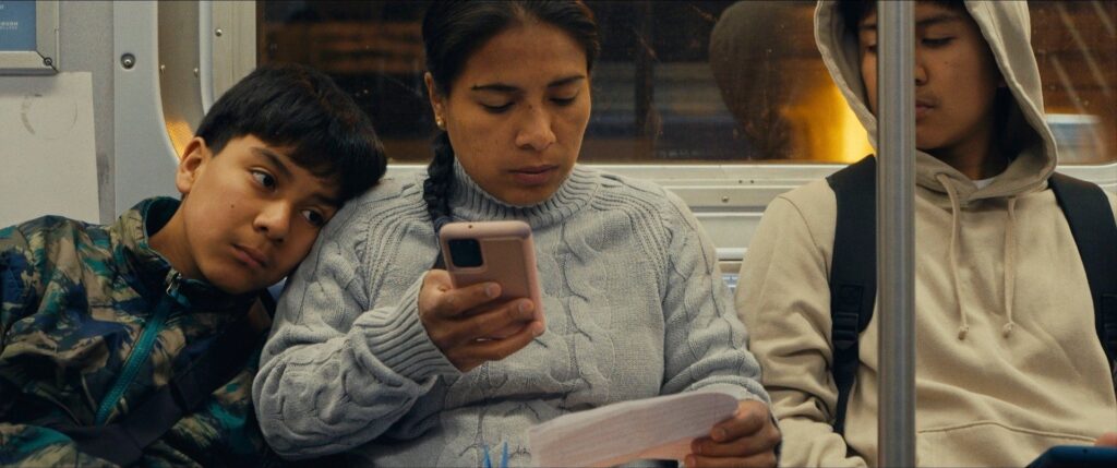 A Latin American woman rides the subway with her two sons. She is sitting between them reading a piece of paper and holding a phone.