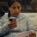 A Latin American woman rides the subway with her two sons. She is sitting between them reading a piece of paper and holding a phone.