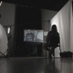 A silhouette of a man sitting in a chair. He faces a TV screen, which shows a close-up of a man's face. The man is sitting in a dimly lit room and there is a white curtain on the right.