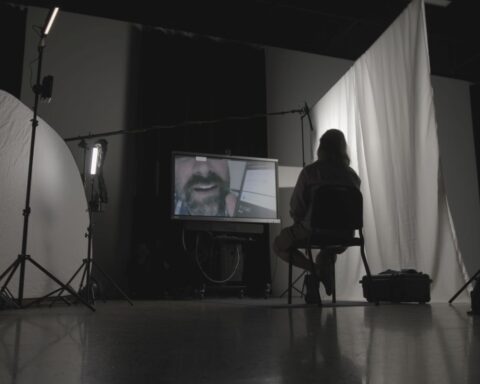 A silhouette of a man sitting in a chair. He faces a TV screen, which shows a close-up of a man's face. The man is sitting in a dimly lit room and there is a white curtain on the right.