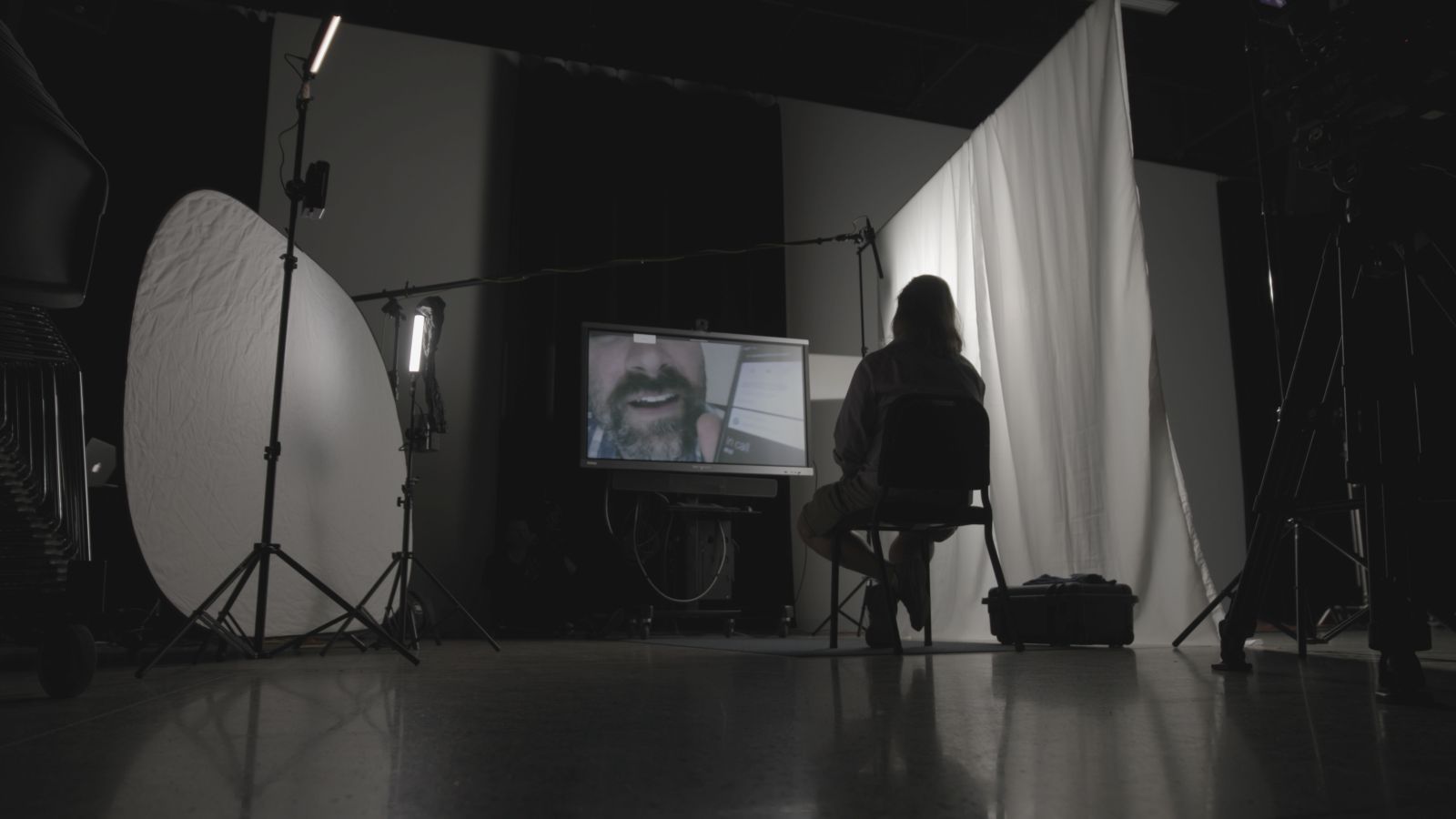 A silhouette of a man sitting in a chair. He faces a TV screen, which shows a close-up of a man's face. The man is sitting in a dimly lit room and there is a white curtain on the right.