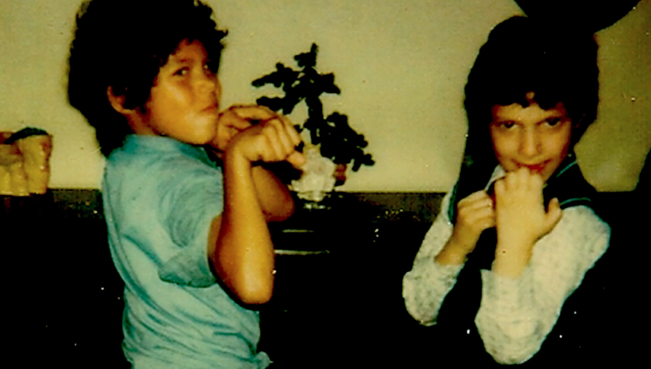 An old photograph of two young boys playing. They have their fists raised and are in a living room.