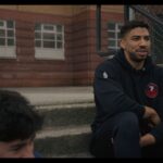 An Indigenous basketball player sits on concrete steps. He is wearing a dark tracksuit.