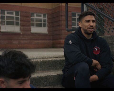 An Indigenous basketball player sits on concrete steps. He is wearing a dark tracksuit.