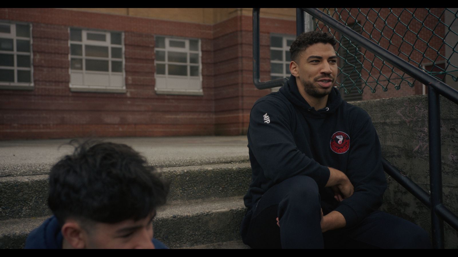 An Indigenous basketball player sits on concrete steps. He is wearing a dark tracksuit.