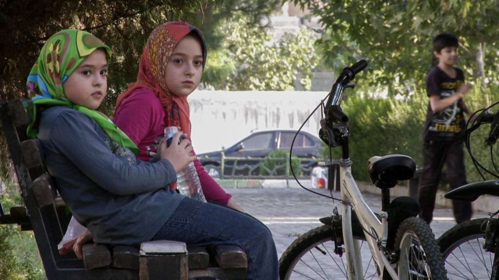 Two girls sit on a bench in an urban park. The bench is on the left of the image and the girls are looking towards the camera.