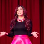 A woman in a black shirt and pink skirt gives a speech. She is facing the camera and has an animated expression with her hands. There is a red curtain behind her.