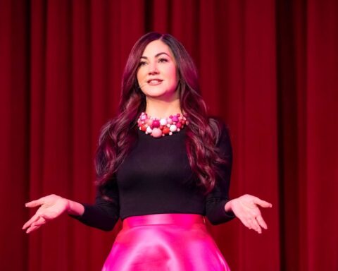 A woman in a black shirt and pink skirt gives a speech. She is facing the camera and has an animated expression with her hands. There is a red curtain behind her.
