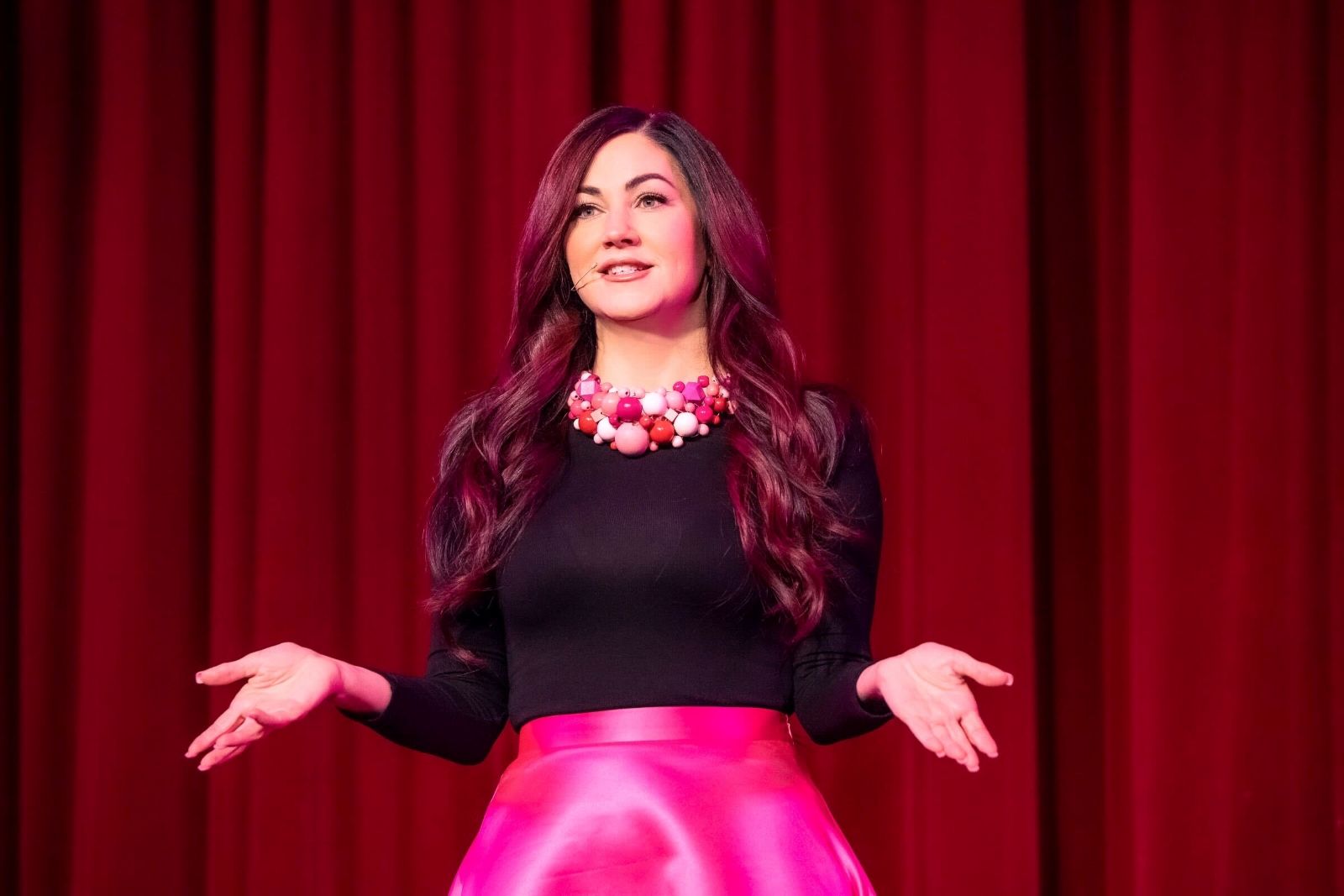 A woman in a black shirt and pink skirt gives a speech. She is facing the camera and has an animated expression with her hands. There is a red curtain behind her.