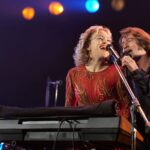 A man and a woman sing sit together at a keyboard and sing into a microphone on stage. She is on the left and isin a red dress with her hair like Patsy Cline, and he is on the right and is dressed up to resemble Neil Diamond.