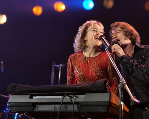A man and a woman sing sit together at a keyboard and sing into a microphone on stage. She is on the left and isin a red dress with her hair like Patsy Cline, and he is on the right and is dressed up to resemble Neil Diamond.