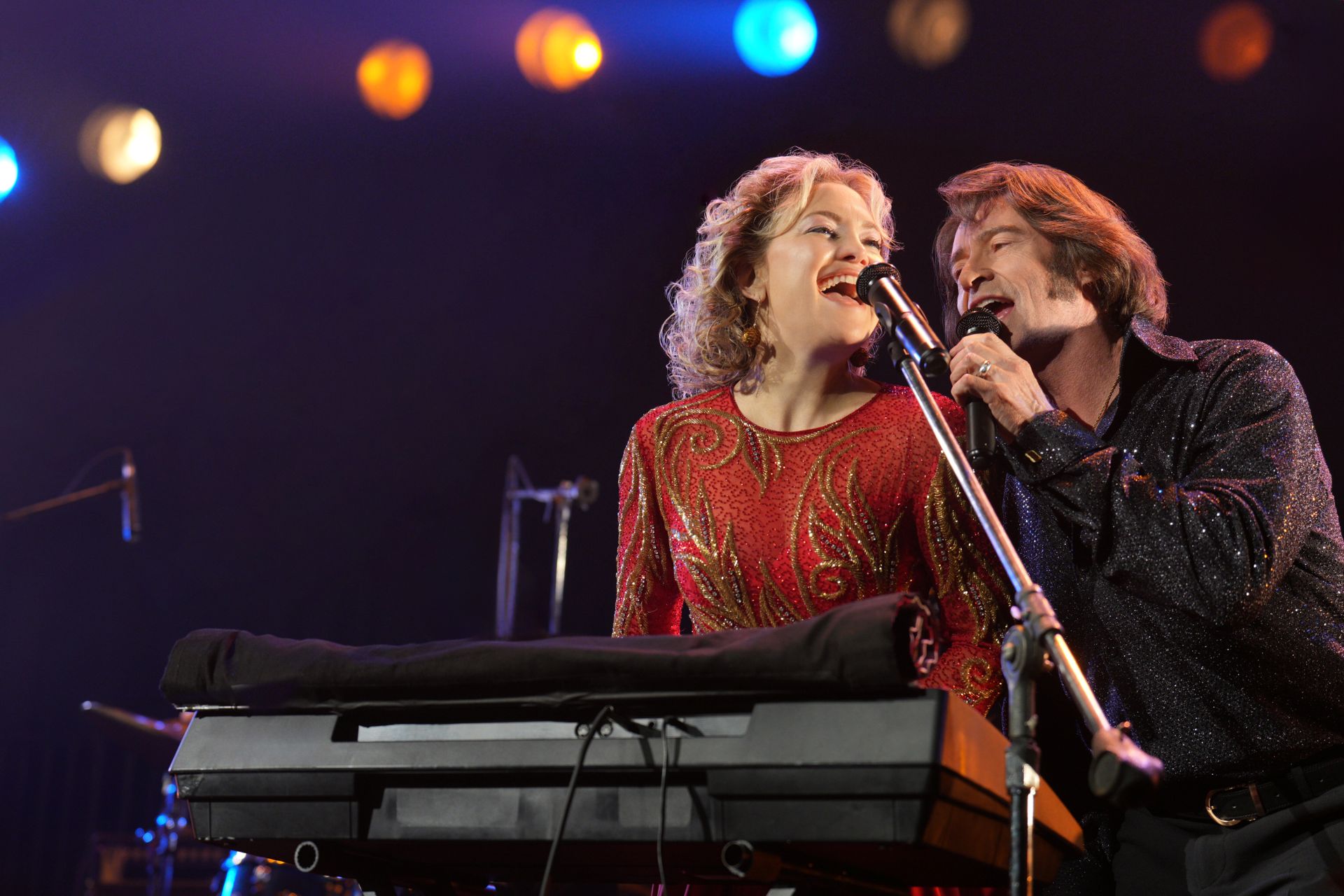 A man and a woman sing sit together at a keyboard and sing into a microphone on stage. She is on the left and isin a red dress with her hair like Patsy Cline, and he is on the right and is dressed up to resemble Neil Diamond.