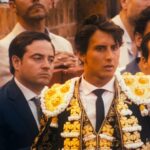 A matador ina black costume adorned with white and gold trim looks out in the bull ring. A crowd watches behind him.