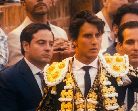 A matador ina black costume adorned with white and gold trim looks out in the bull ring. A crowd watches behind him.