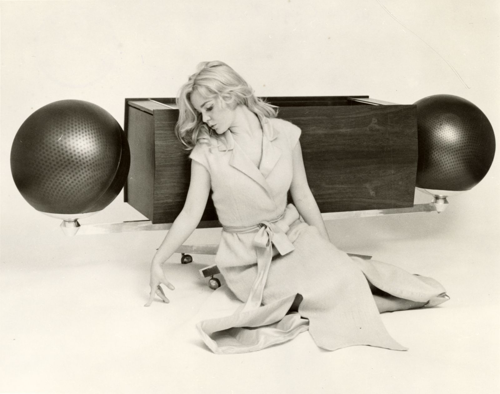 A black and white archival image of a woman sitting in front of a Clairtone stereo.She is reading a magazine while listening to music. There backdrop and floor are white.