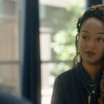 Amanda Parris is seated facing an off-screen interviewee. She is a Black woman with blond braided hair, and is wearing a dark blue collared shirt.