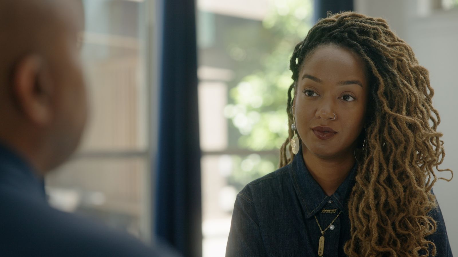 Amanda Parris is seated facing an off-screen interviewee. She is a Black woman with blond braided hair, and is wearing a dark blue collared shirt.