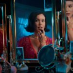 Andrea Werhun is a white woman with brown hair, wearing a pink dress with a V-neckline. She is sitting down at a make-up table, looking at a three-paned mirror and applying lipstick.