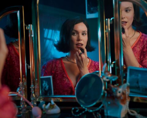Andrea Werhun is a white woman with brown hair, wearing a pink dress with a V-neckline. She is sitting down at a make-up table, looking at a three-paned mirror and applying lipstick.