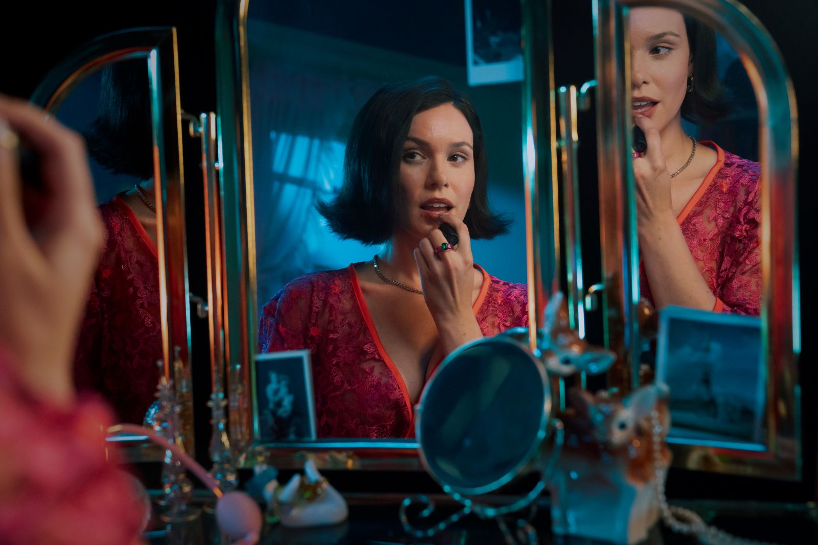 Andrea Werhun is a white woman with brown hair, wearing a pink dress with a V-neckline. She is sitting down at a make-up table, looking at a three-paned mirror and applying lipstick.