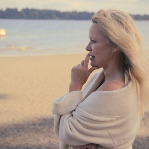 Actress Pamela Anderson stands on a beach and looks reflectively at the water. Her hand is raised to her mouth. She is wearing a white sweater.