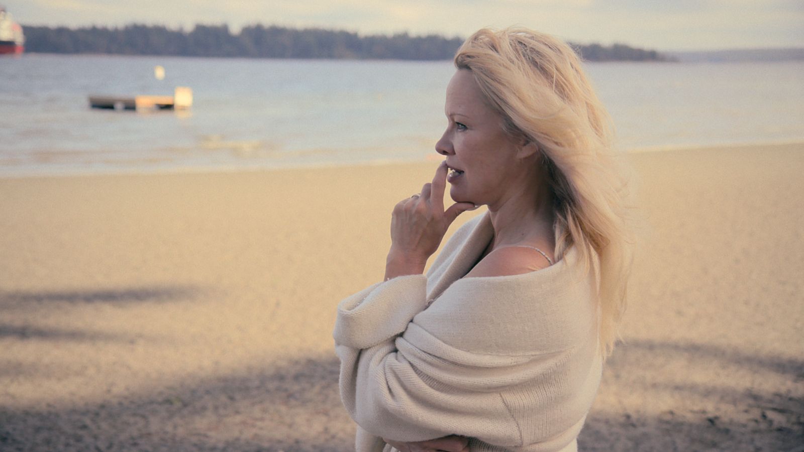 Actress Pamela Anderson stands on a beach and looks reflectively at the water. Her hand is raised to her mouth. She is wearing a white sweater.