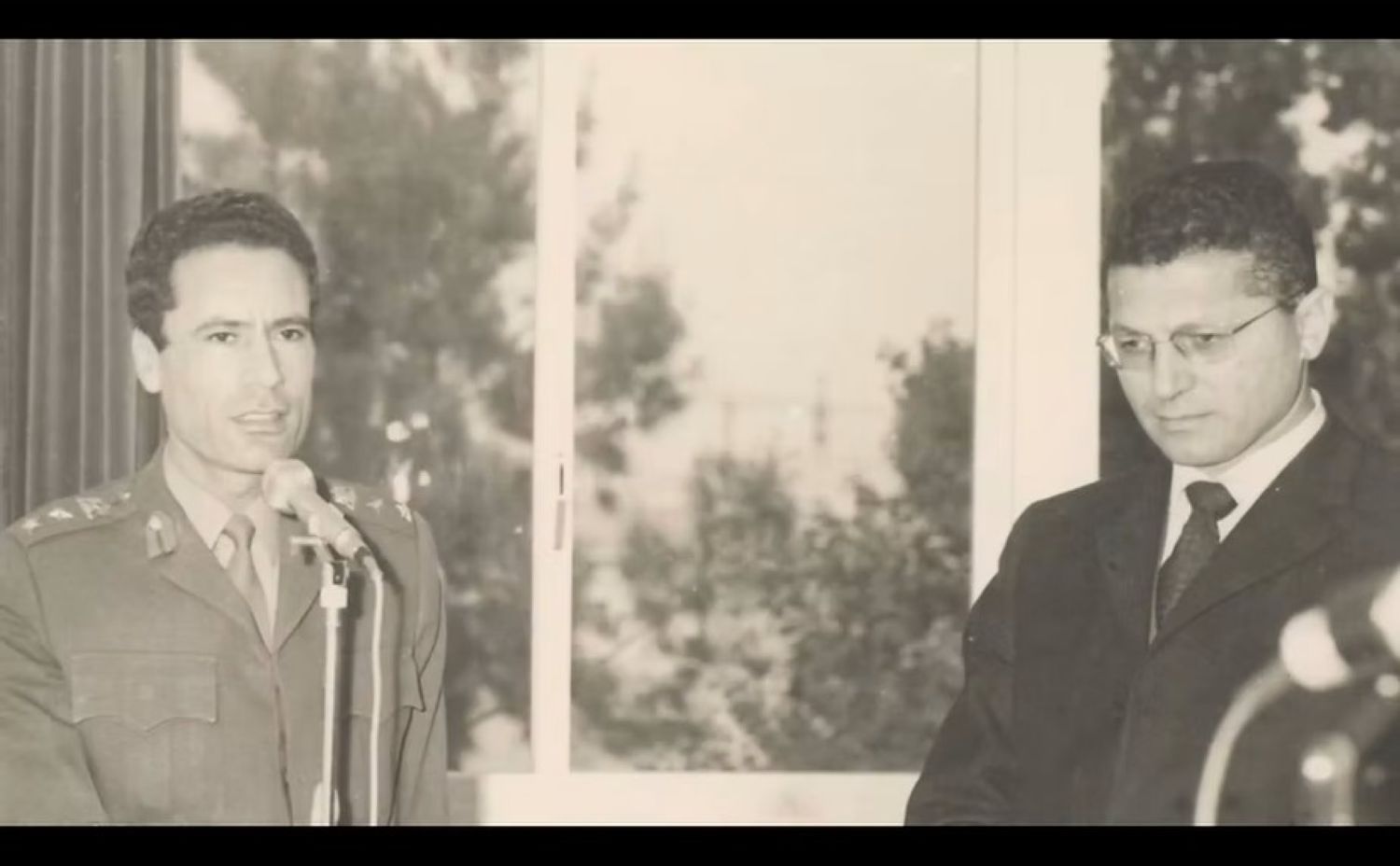 An archival black and white image of a young Muammar Gaddafi standing with a man in a suit.