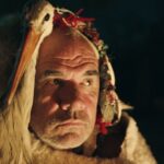 An older man and and a white stork sit by the fire as a group performs a traditional Macedonian ritual.