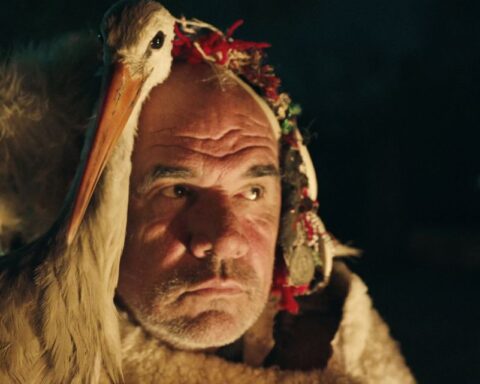 An older man and and a white stork sit by the fire as a group performs a traditional Macedonian ritual.