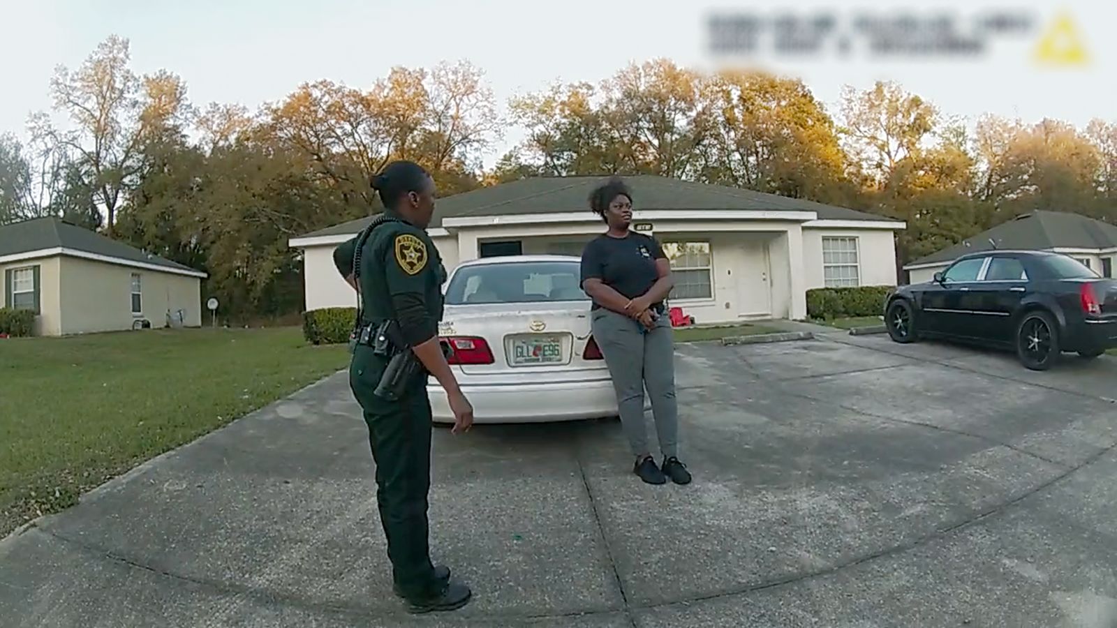 Two Black women, one police officer and one civilian, stand in a driveway having a conversation. They are in front of a white car and bungalow. They are seen through the curved view of a body cam affixed to another police officer.