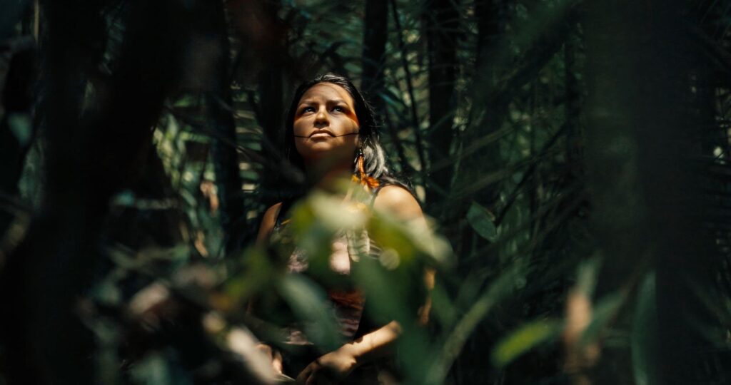 An Indigenous woman stands amid the trees of the Amazon. She looks up to the tree tops as sunlight brightens her face.