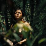 An Indigenous woman stands amid the trees of the Amazon. She looks up to the tree tops as sunlight brightens her face.