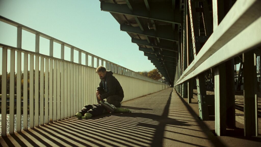 A man bends down beside the railing of a bridge, examining a dummy shaped like a human body.
