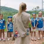 Five Girl Scouts in uniform face their leader, who has her back to the camera.