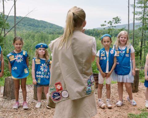 Five Girl Scouts in uniform face their leader, who has her back to the camera.