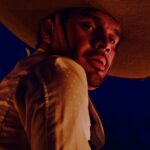 A Mexican cowboy is pictured in close-up as the camera looks upward towards his face. He is looking downward and wearing a large sombrero. It is nighttime.