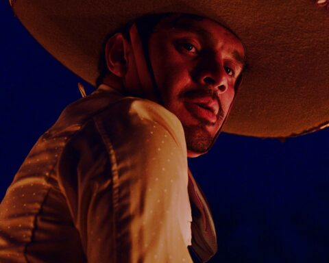 A Mexican cowboy is pictured in close-up as the camera looks upward towards his face. He is looking downward and wearing a large sombrero. It is nighttime.