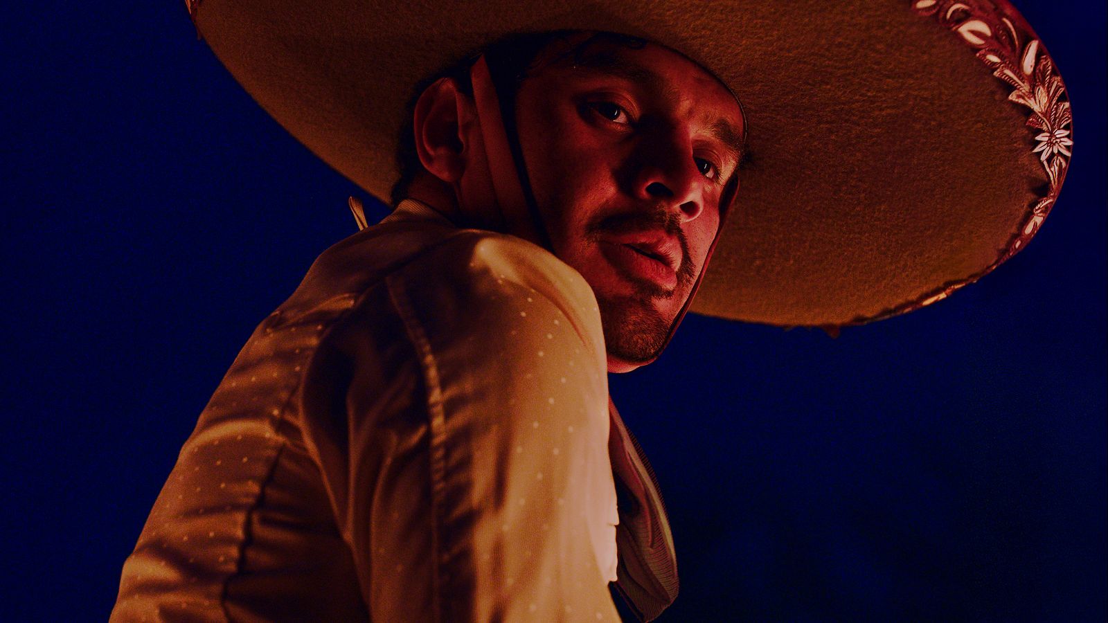 A Mexican cowboy is pictured in close-up as the camera looks upward towards his face. He is looking downward and wearing a large sombrero. It is nighttime.