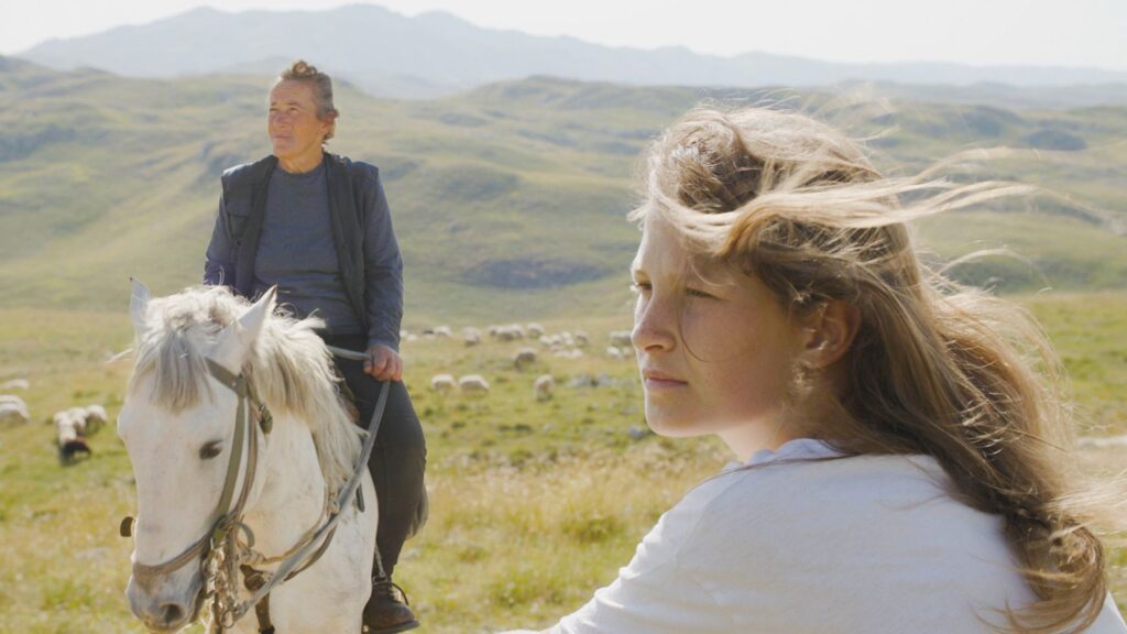 A young woman sits at the foreground of the frame looking off to the left. Her mother is behind her, sitting on a white horse. The rolling pasturelands are in the background.