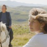 A young woman sits at the foreground of the frame looking off to the left. Her mother is behind her, sitting on a white horse. The rolling pasturelands are in the background.
